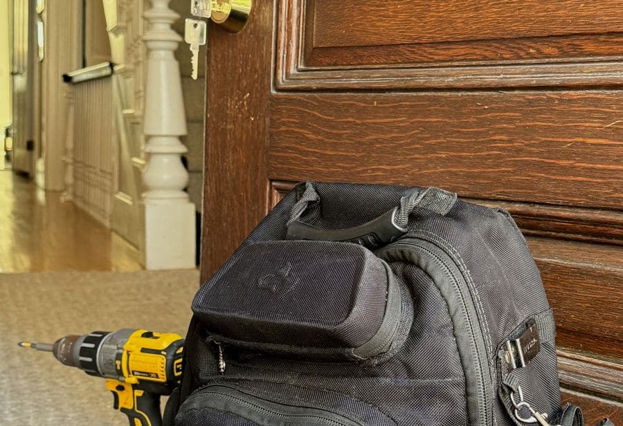 Tool backpack and cordless drill on the floor in front of a dark wood door with a brass deadbolt